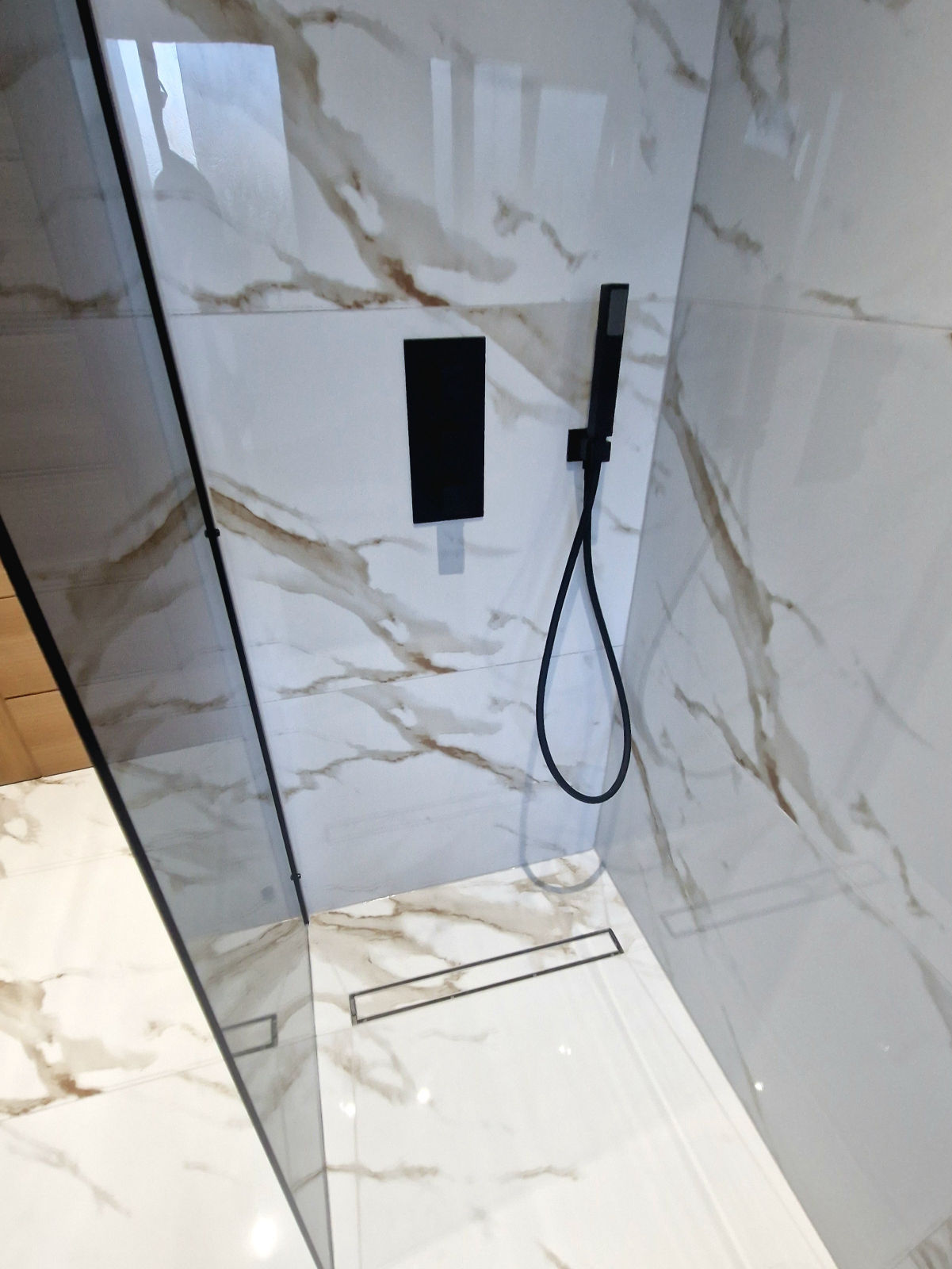 A marble walk-in shower with concealed black valves, a hand shower and a linear drain set into the floor