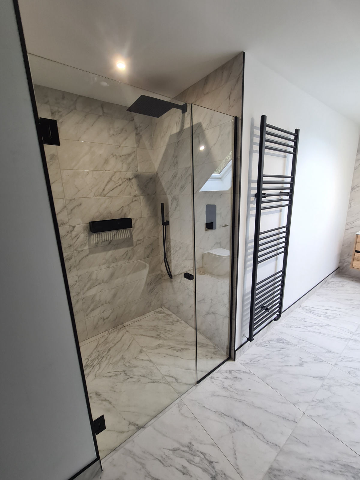 A walk-in shower with a black rainfall head, a recessed shelf and a black ladder towel rail