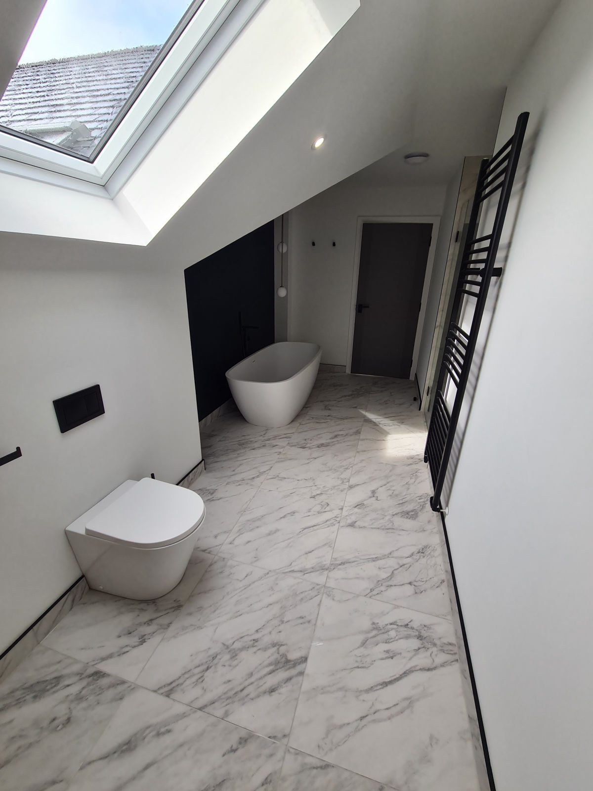 A loft bathroom with a freestanding bath against a black panel, under a roof window