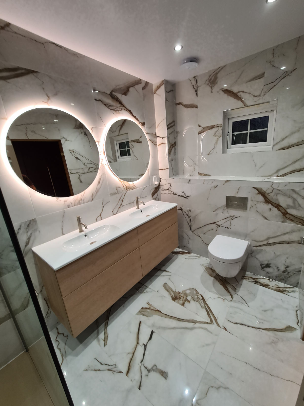 A marble bathroom with twin backlit round mirrors above an oak double vanity