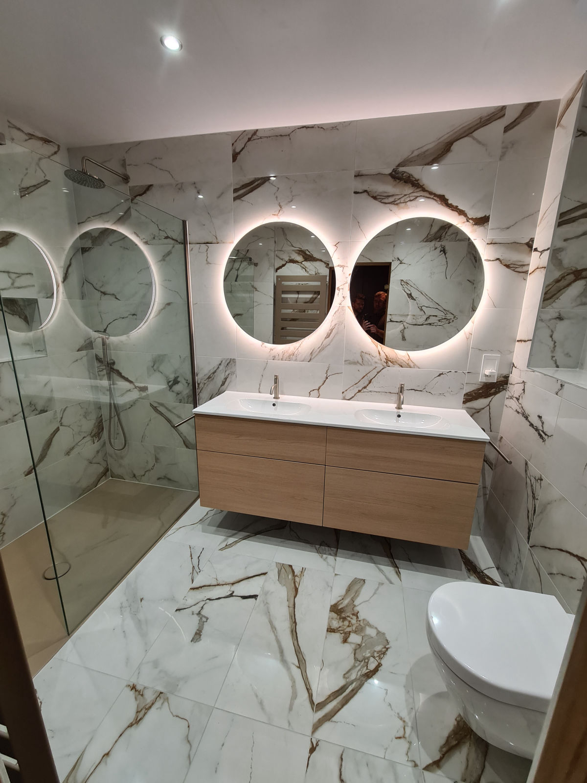 A marble-tiled bathroom with twin backlit round mirrors, an oak double vanity and a walk-in shower